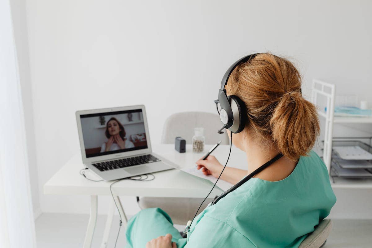 A healthcare professional wearing headphones sits at a desk and conducts a virtual consultation with a patient on a laptop.