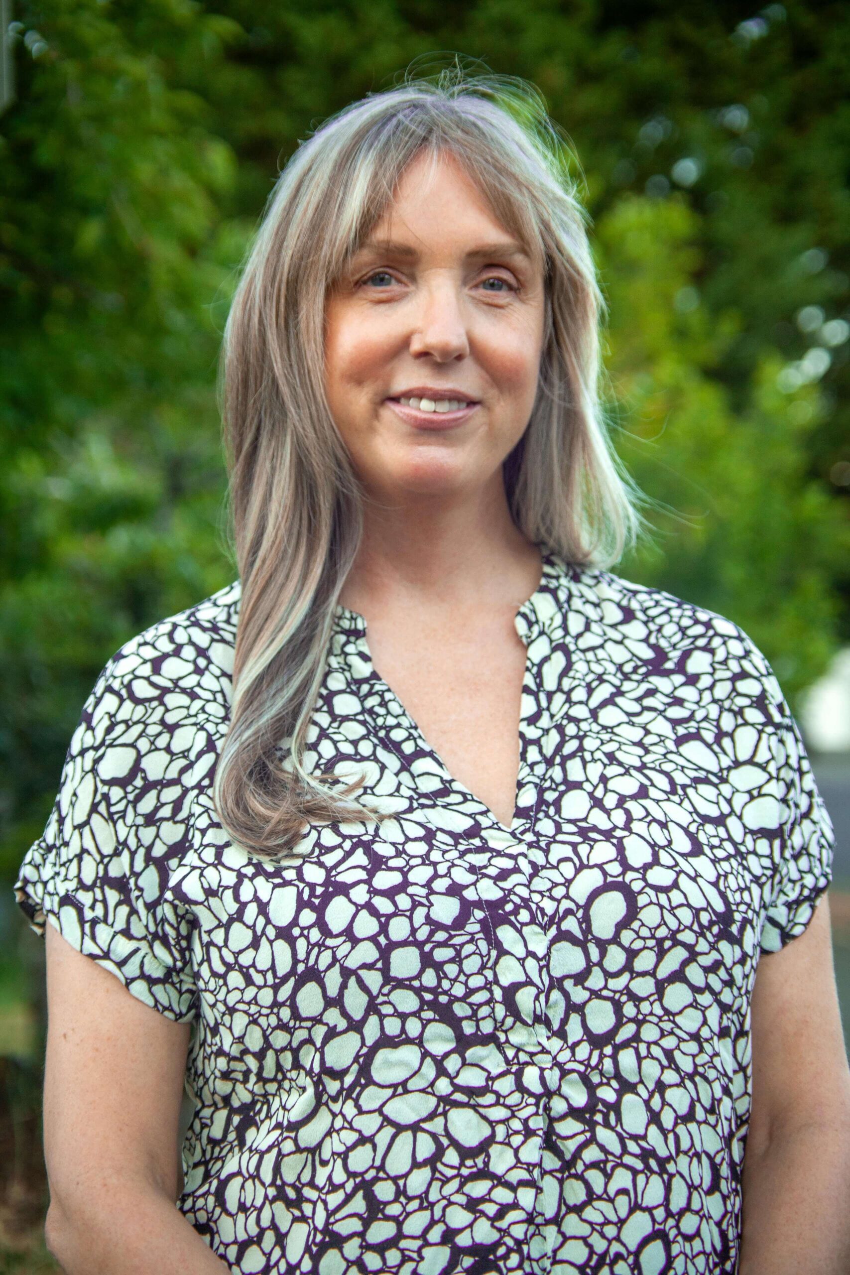A woman with long, light brown hair stands outdoors, wearing a short-sleeved, patterned blouse. Green foliage is visible in the background.