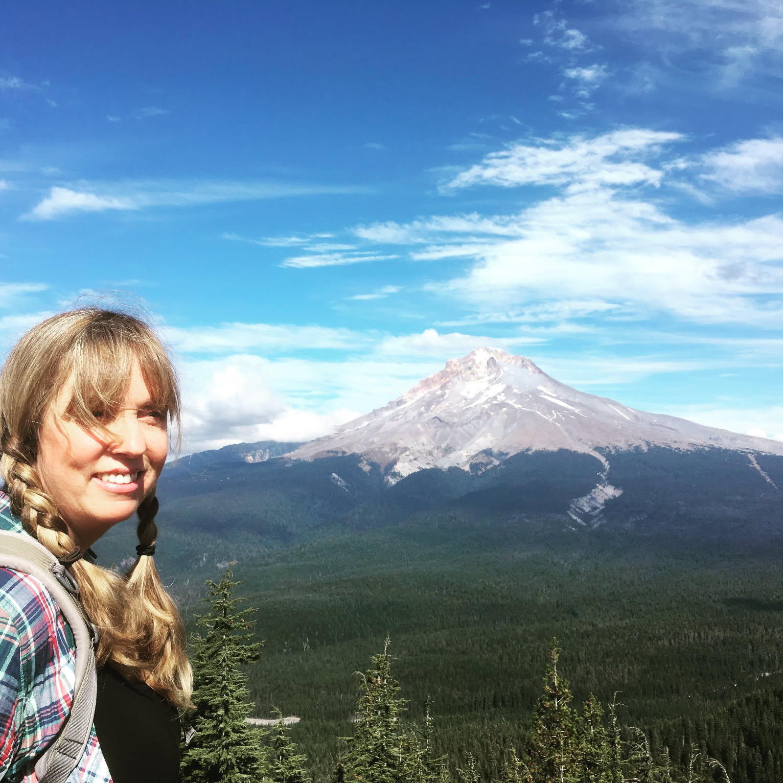 A woman with blonde braids and a backpack stands in front of a forested landscape with a snow-capped mountain under a blue sky.