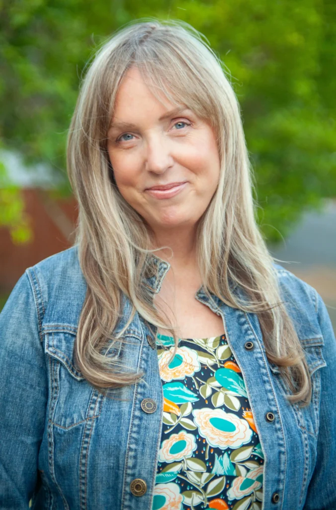 A woman with long blonde hair, wearing a denim jacket over a floral top, stands outdoors with greenery in the background.